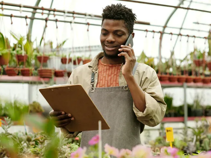 Garden center owner on the phone with a clipboard among hanging plants and flowers, Town Directory Base